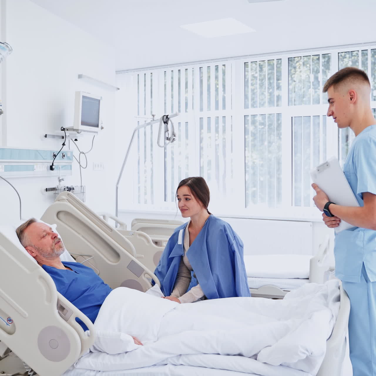 Young doctor talks to a patient. Senior man laying in hospital bed and his daughter visiting him on the modern clinic background.