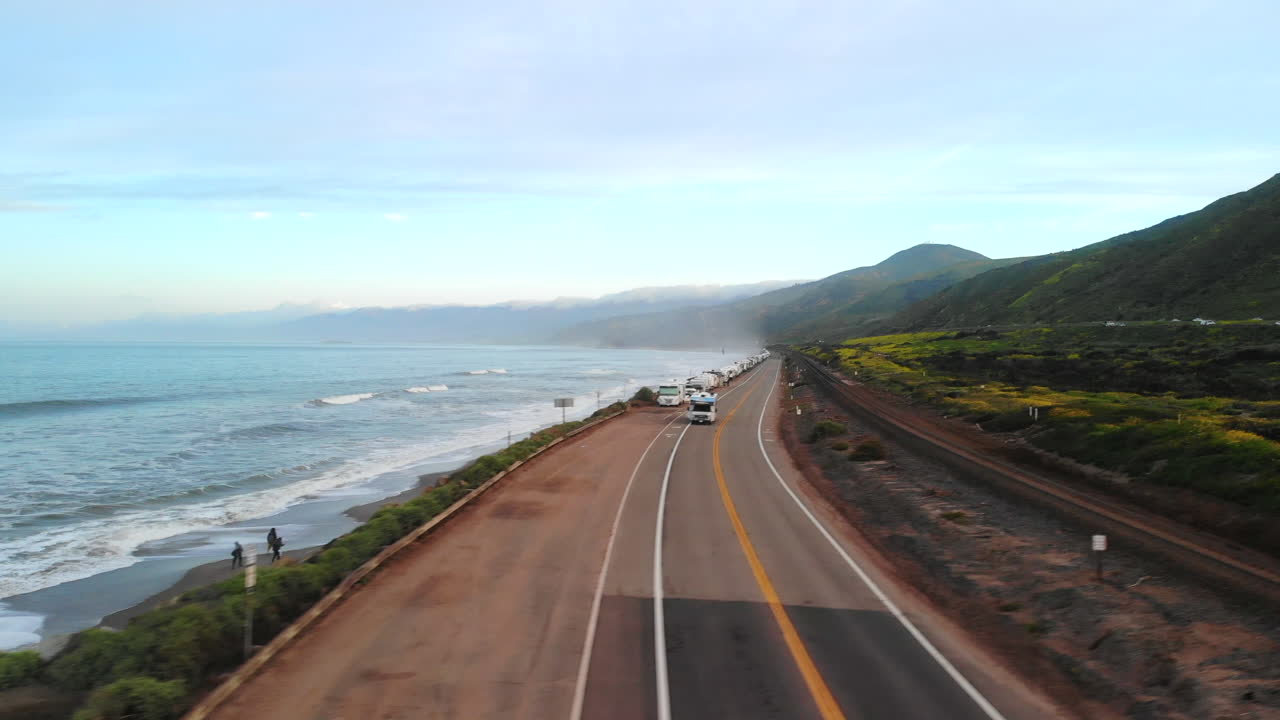 Aerial drone flying low over the ocean waves and rising up over the beach coastline at sunrise with people and rv vehicles camping on a road in a Ventura, California