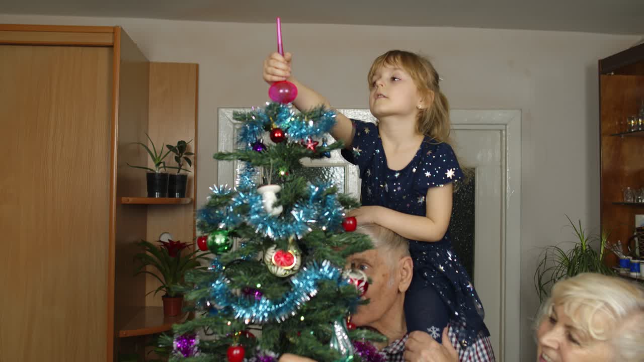 Children girl with elderly couple grandparents decorating artificial Christmas pine tree at home