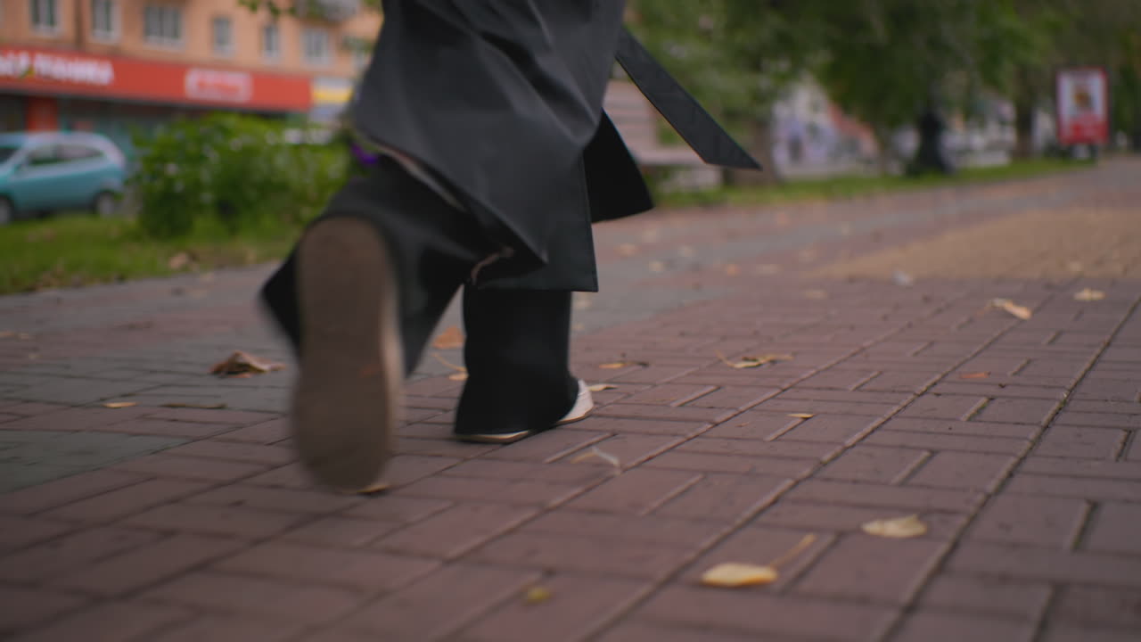 Lower shot of woman running along brick sidewalk in autumn city, long black trench coat flowing as feet step forward, dry fallen leaves scattered on pavement creating seasonal urban atmosphere