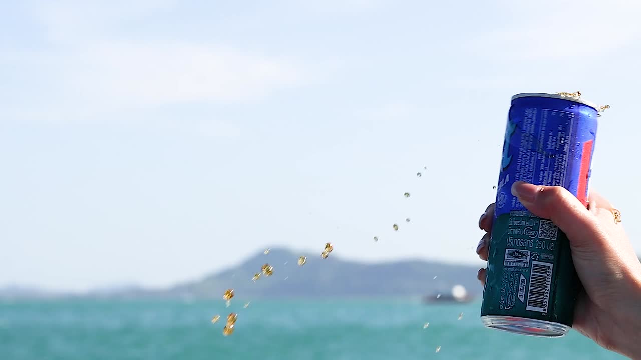 A hand shakes a blue beverage can causing liquid to spray against a tropical seascape