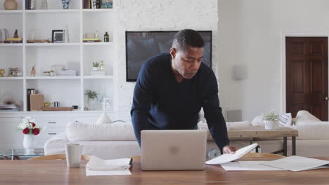 Middle-aged-man-standing-at-dining-room-table-looking-at-paperwork-and-using-laptop-computer