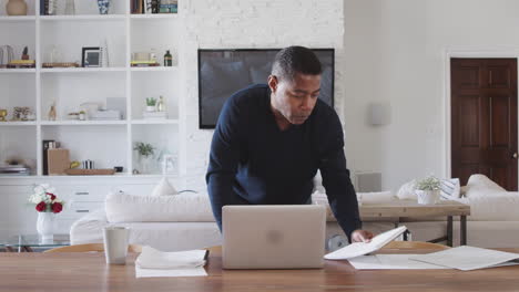 Un-Hombre-De-Mediana-Edad-Parado-En-La-Mesa-Del-Comedor-Mirando-El-Papeleo-Y-Usando-Una-Computadora-Portátil