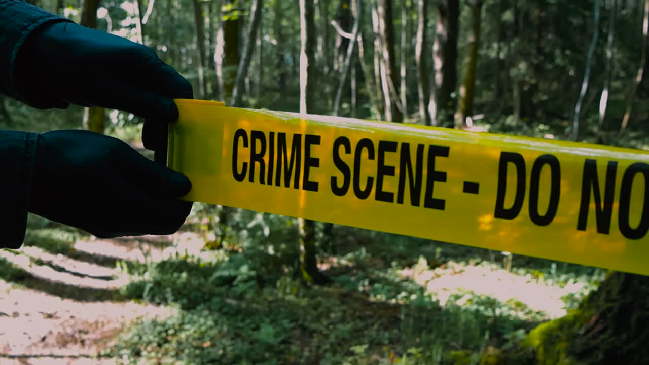 Close up footage of a investigator with black nylon gloves unrolling or unraveling a Crime Scene yellow colored police caution tape in front of a sunny forest hiking road during sunny day in slow-mo