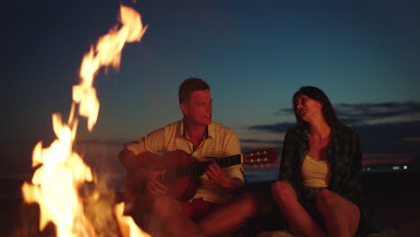una pareja tocando la guitarra junto a una fogata en la playa al atardecer