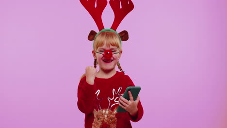 niña con cuernos de ciervo de navidad mirando pantalla de teléfono inteligente sinceramente regocijándose ganar éxito suerte