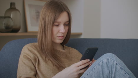 Young-woman-sitting-at-desk-at-home-office-distracted-from-work-holding-smart-phone-using-social-networking-website-chatting-with-friend-remotely-using-modern-device-and-internet-connection