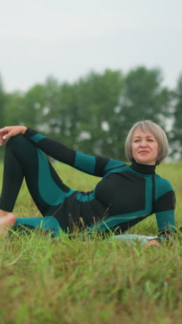 middle aged woman lying on her side with hand resting on her leg, looking thoughtfully into the distance, surrounded by greenery and trees in the background