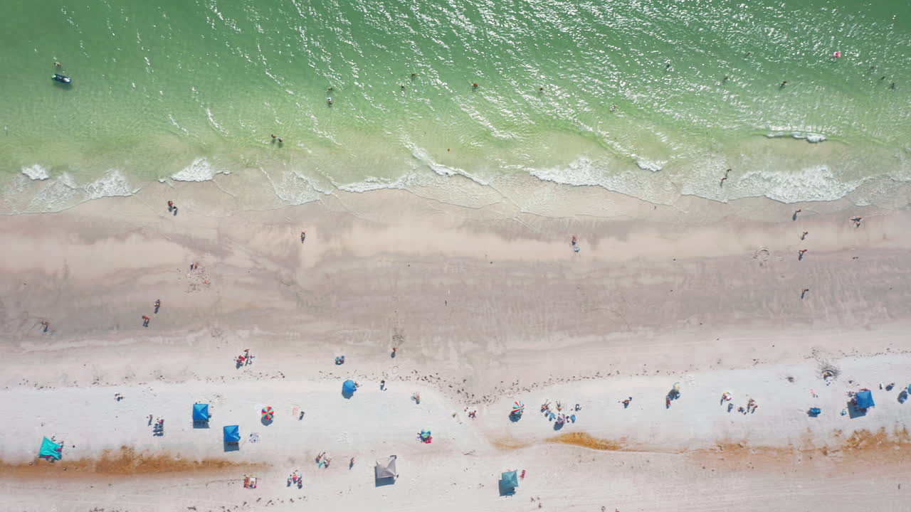 Turquoise ocean waves meet the sandy shoreline as beachgoers relax beneath colorful umbrellas, captured from a top-down aerial perspective highlighting coastal textures and colors