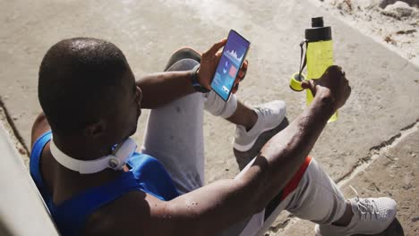 Deportista-Afroamericano-Sentado-En-La-Acera,-Usando-Un-Teléfono-Inteligente-Con-Estadísticas-En-Pantalla