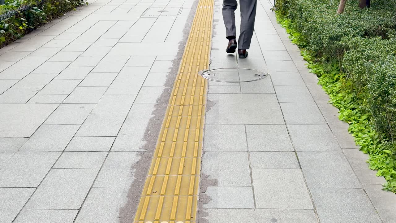 Low angle shot follows a businessman walking on a sidewalk with yellow tactile paving