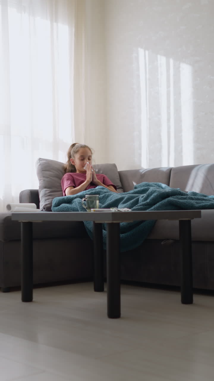 Ragazza con l'influenza sdraiata sul divano, avvolta in una coperta, che starnutisce in un fazzoletto, tenendolo al naso, che si riposa mentre guarisce dalla malattia con la luce del sole che filtra dalla finestra