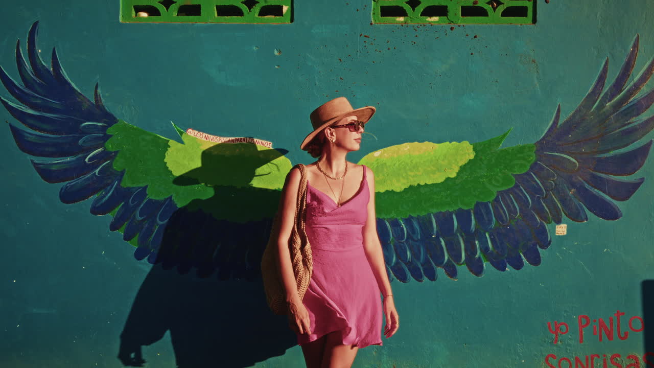 Beautiful young girl with a pink dress and a hat standing in from of a turquoise wall on which are painted some parrot wings
