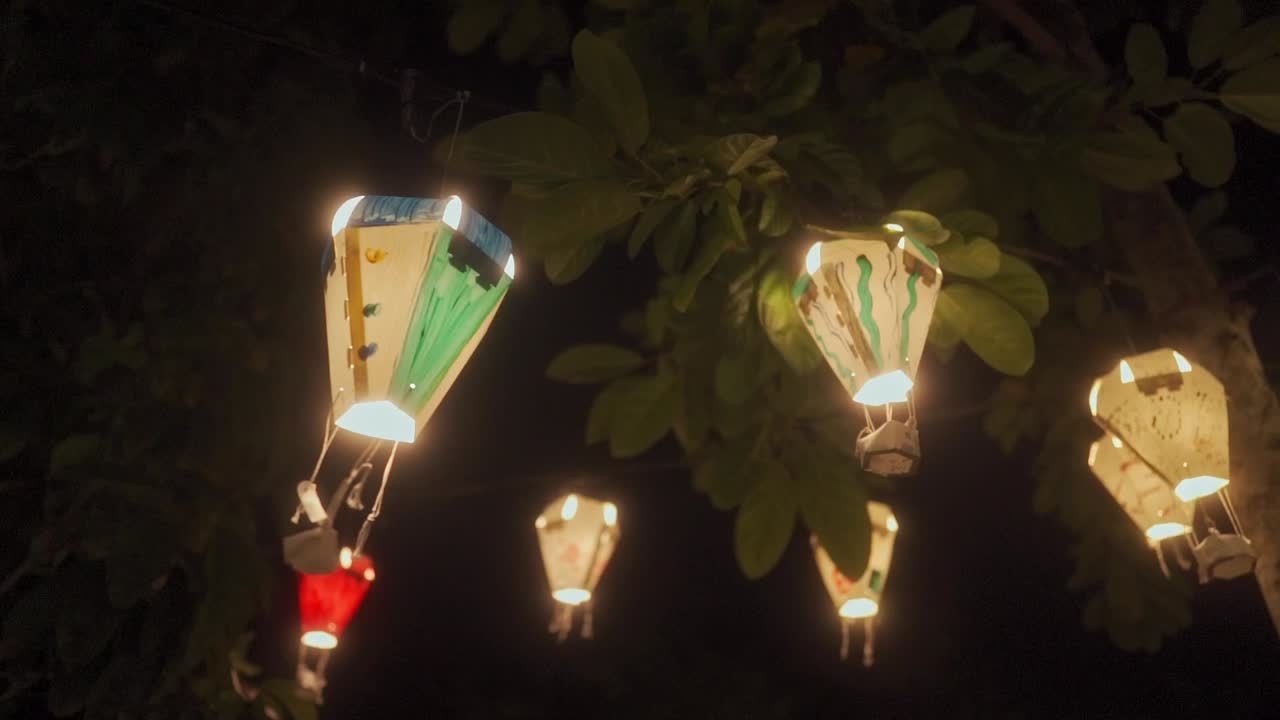 Serangkaian lampion kertas dekoratif yang dilukis tangan berbentuk seperti balon udara panas bersinar hangat saat tergantung dari dahan pohon di malam hari.