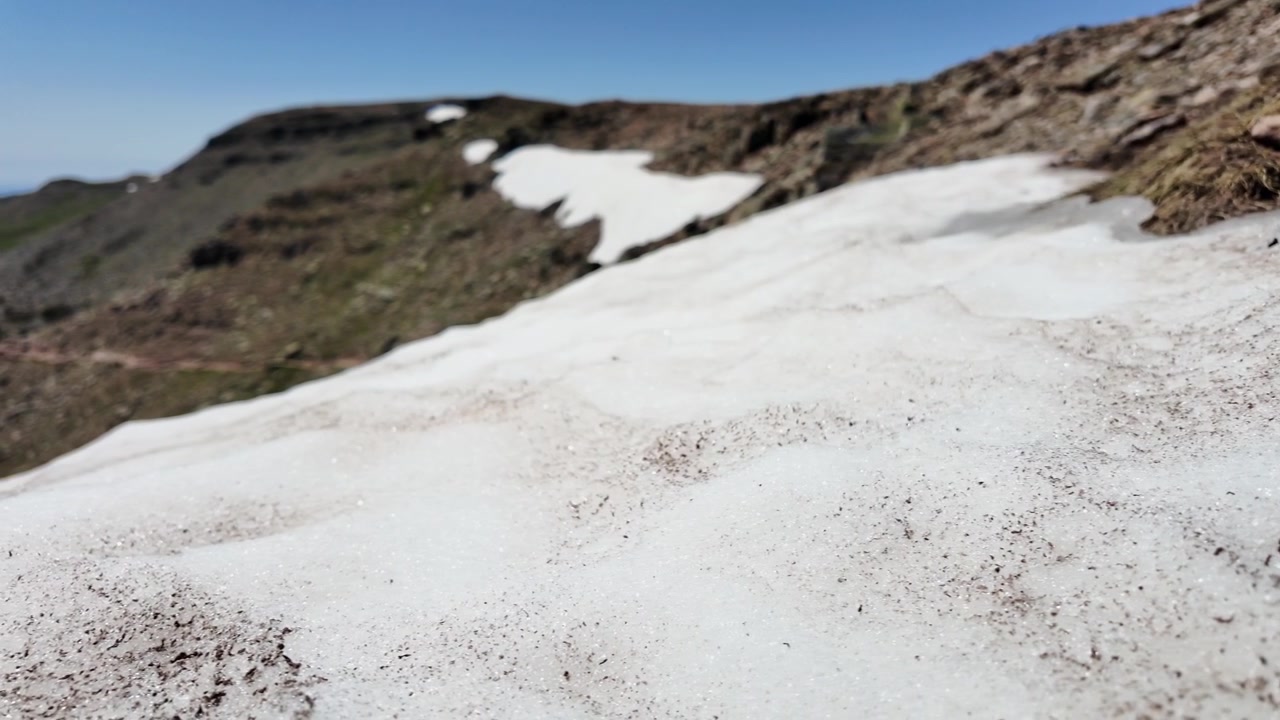 Ripresa panoramica a scorrimento di paesaggio montano mostra cime innevate che si sciolgono sulle aspre montagne spagnole sotto un cielo azzurro e sereno, evidenziando lo scioglimento precoce dovuto al riscaldamento globale. Luce diurna intensa, grandangolo statico