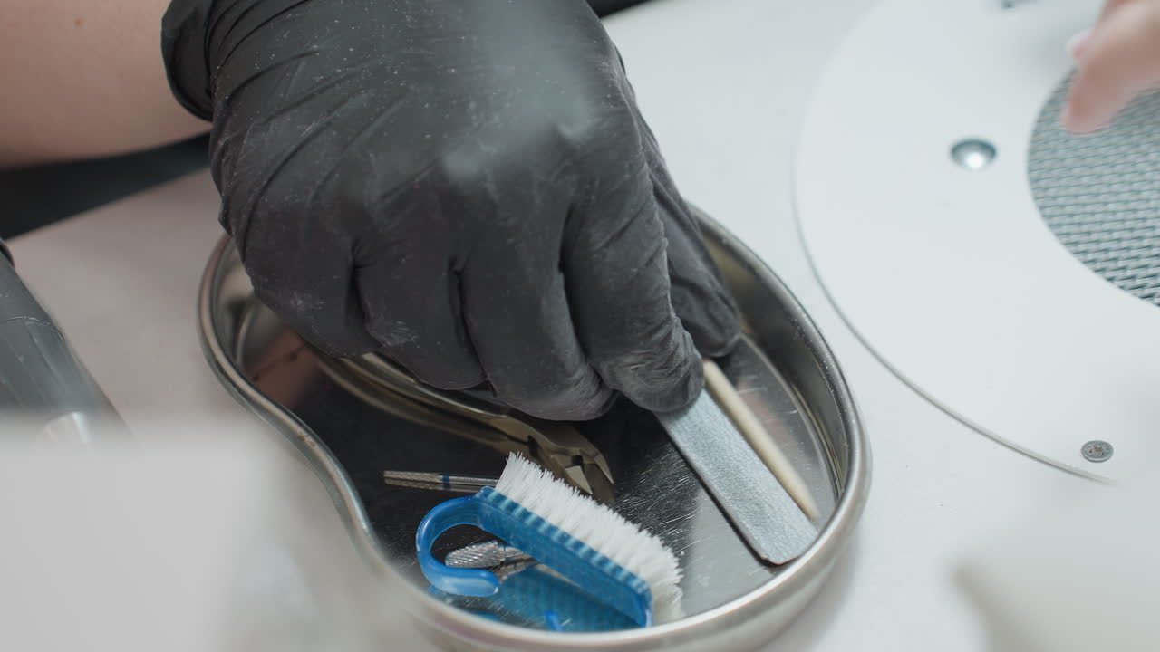 Close-up of nail technician wearing black glove selecting manicure tool from stainless tray containing various nail care instruments including buffer, stick, brush and clipper beside customer hand