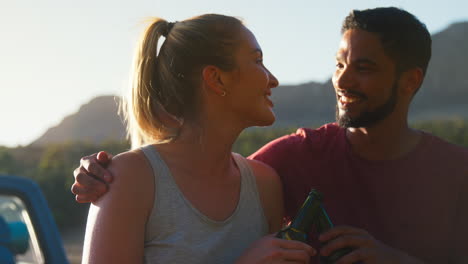 couple with backpacks sitting on tailgate of pick up truck on road trip by lake drinking beer