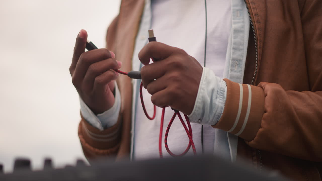 black sound engineer linking red cable into mixer on rooftop, methodical hands threading jack, overcast skyline and urban backdrop, warm jacket sleeves, tactile checks before live outdoor set