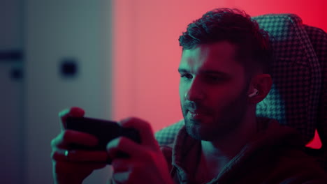 jugador adicto jugando por teléfono inteligente nueva aplicación con videojuegos retrato de joven emocional en casa en la noche escuchando sonido por auriculares en el oído