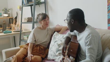 multiethnic couple playing guitar and singing on sofa at home