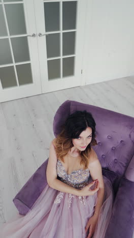 attractive girl in a sexy dress sitting in a purple armchair. beauty gorgeous bride is posing in a nice interior room. beautiful model in wedding dress. camera moves up. vertical video