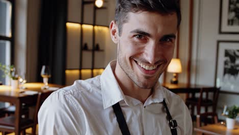 young charming waiter, wearing a white shirt and black apron, displays a range of emotions, from serious to a genuine smile, within a cozy restaurant setting