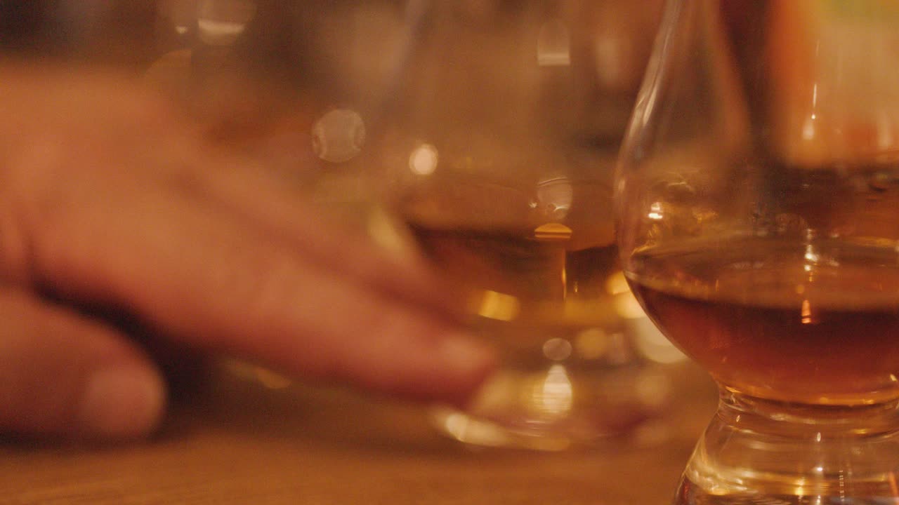 A hand slowly reaches for and lifts a whisky glass from a rustic wooden table, under soft, warm ambient lighting, with shallow depth of field