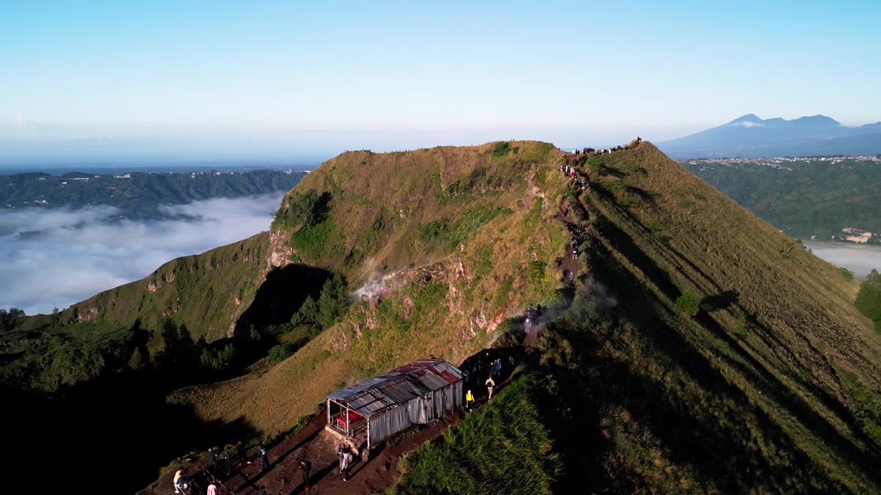 Escursionisti sul Monte Batur a Bali che camminano su uno stretto sentiero di cresta lungo il bordo del cratere con fumo e vapore vulcanico visibili da un vulcano attivo in un terreno ad alta quota sopra le nuvole, riprese aeree con drone ad arco.