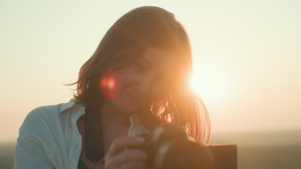 Close up of smiling woman holding camera while reviewing shots during golden hour, soft sun flare glowing from behind her hair, relaxed moment captured in peaceful natural landscape