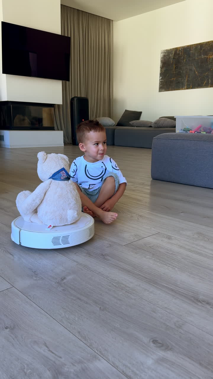 Lovely kid sits on the floor next to the robot cleaner with teddy sitting on it. Excited baby boy watches the device move by the room. Vertical video.