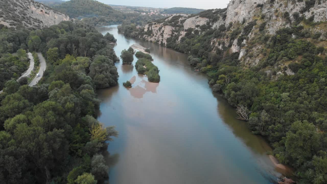 Video drone in volo sul fiume Nestos in Grecia, rocce e scogliere sulla destra e alberi e cespugli sulla sinistra, un villaggio in lontananza in una giornata estiva.