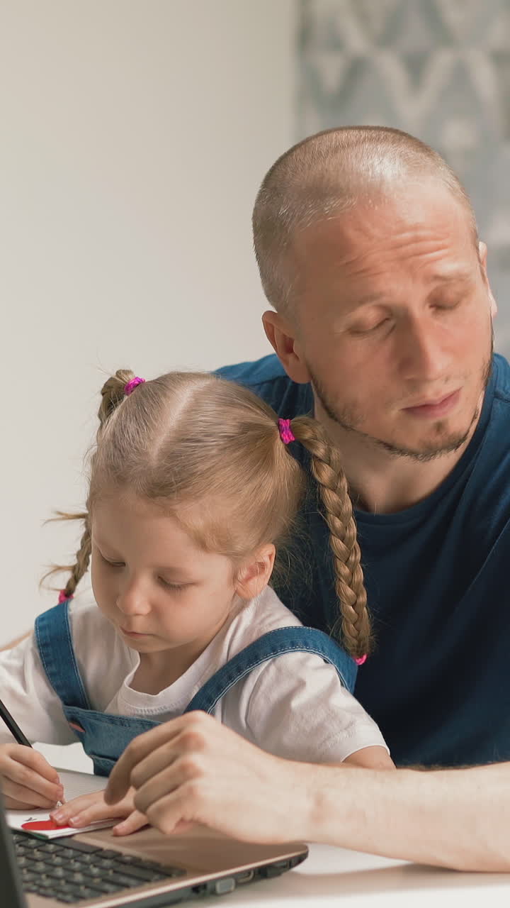 concept of: little daughter sits on her dad's lap writes in notebook, listens to teacher from laptop, is on distance learning, father misses, dreamily looks out window, helps