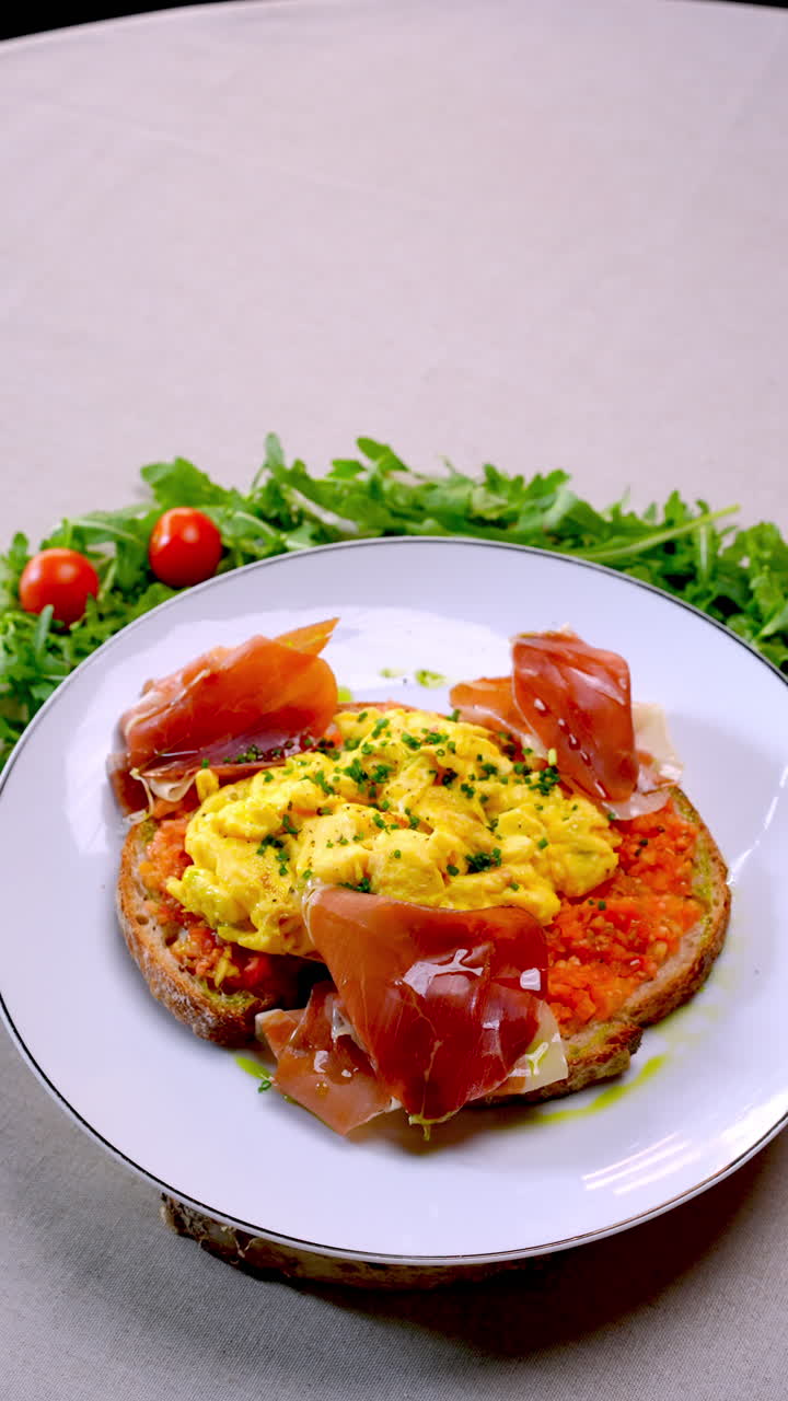 Hearty breakfast plate featuring fluffy scrambled eggs nestled on crispy toasted bread, garnished with savory ham, peppery arugula, vibrant cherry tomatoes, and sprinkled chives