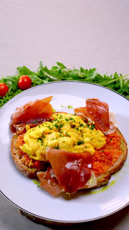 hearty breakfast plate featuring fluffy scrambled eggs nestled on crispy toasted bread, garnished with savory ham, peppery arugula, vibrant cherry tomatoes, and sprinkled chives