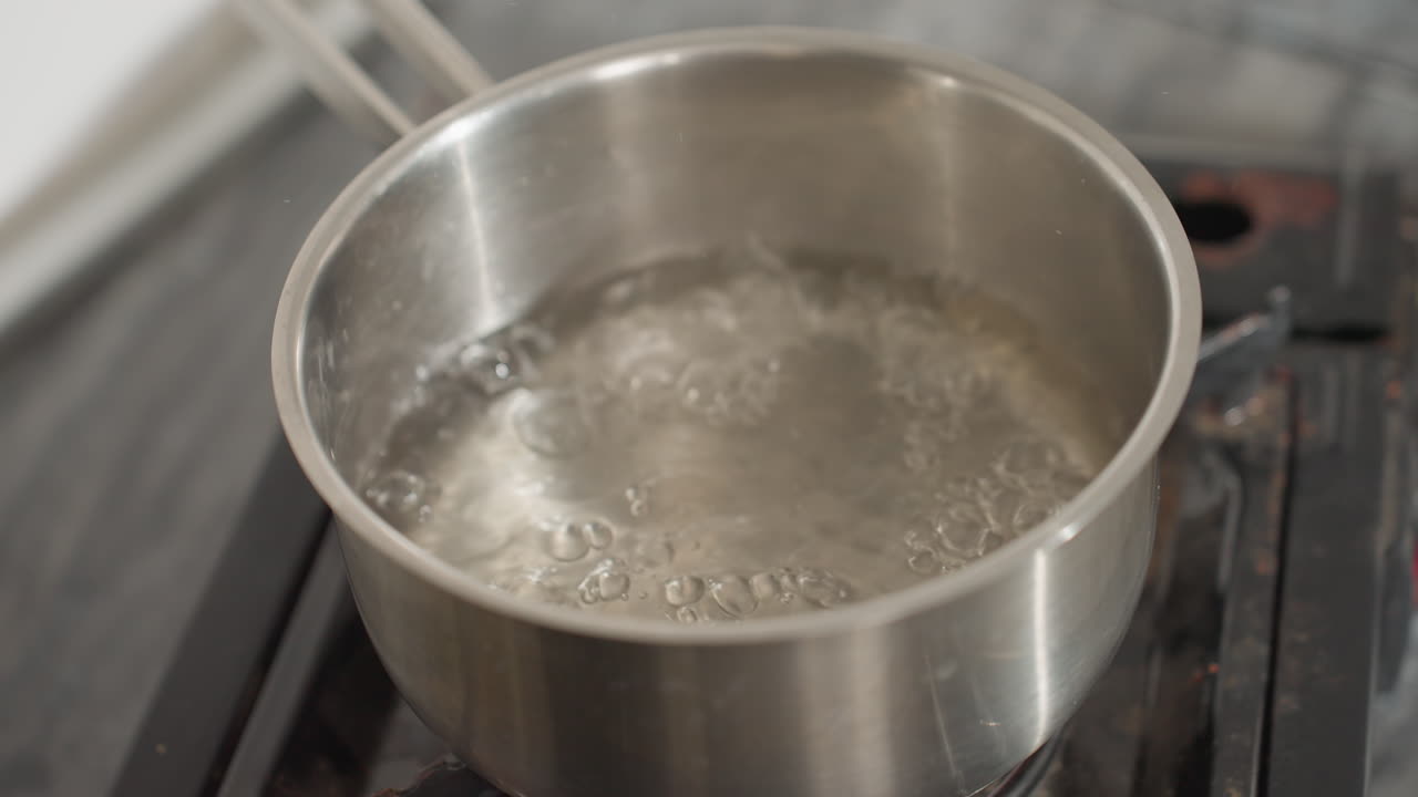 Close up of water steaming and boiling vigorously in stainless steel pot placed on gas burner in kitchen, bubbles rapidly rising to surface, intense heating process visible