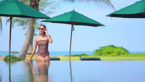 Vacaciones-En-Un-Destino-Tropical-Exótico,-Hermosa-Mujer-Asiática-Sentada-En-El-Borde-De-La-Piscina-Infinita-En-Traje-De-Baño-Con-Follaje-Verde-Y-Mar-De-Fondo,-Cuadro-Completo