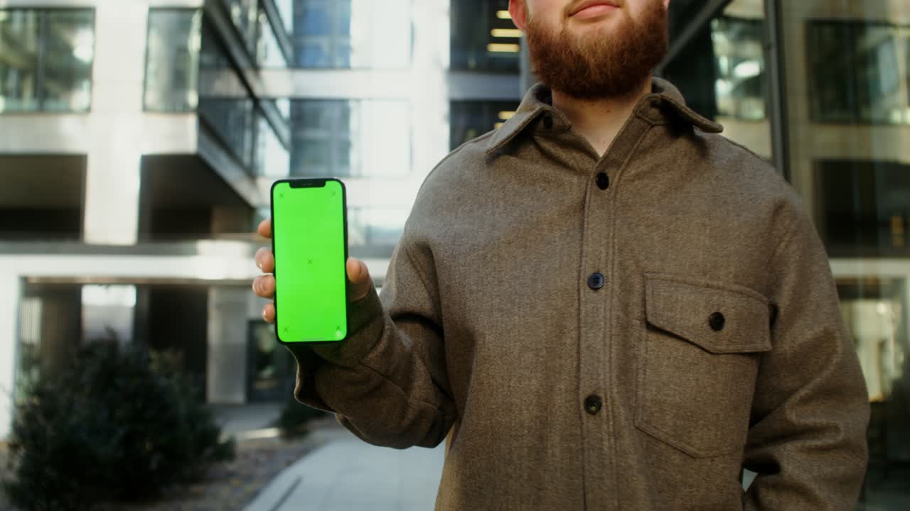 hombre sosteniendo un teléfono inteligente con pantalla verde frente a un edificio moderno