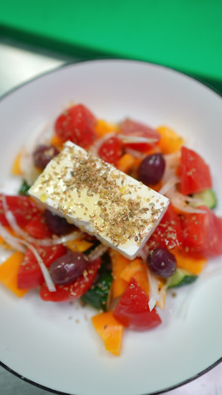 Vertical footage Professional chef wearing gloves carefully sprinkling aromatic oregano over colorful greek salad featuring feta cheese, ripe tomatoes, cucumbers, onions, and kalamata olives