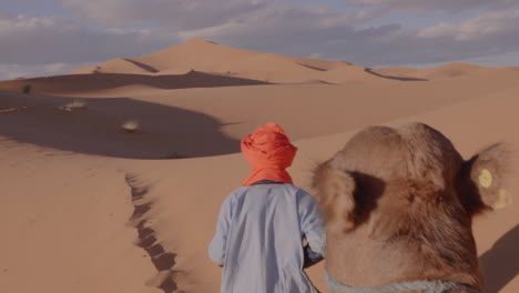 pov riding a camel into the sahara desert, guided by man in turban