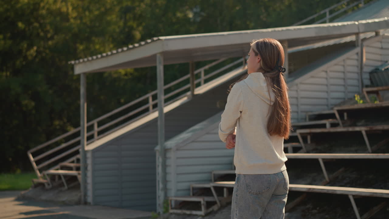 팔을 접고 후디를 입은 젊은 여성이 경기장 좌석 근처의 야외에서 사려 깊게 고 있으며, 나무로 둘러싸인 조용한 길을 따라 움직이고 있습니다.