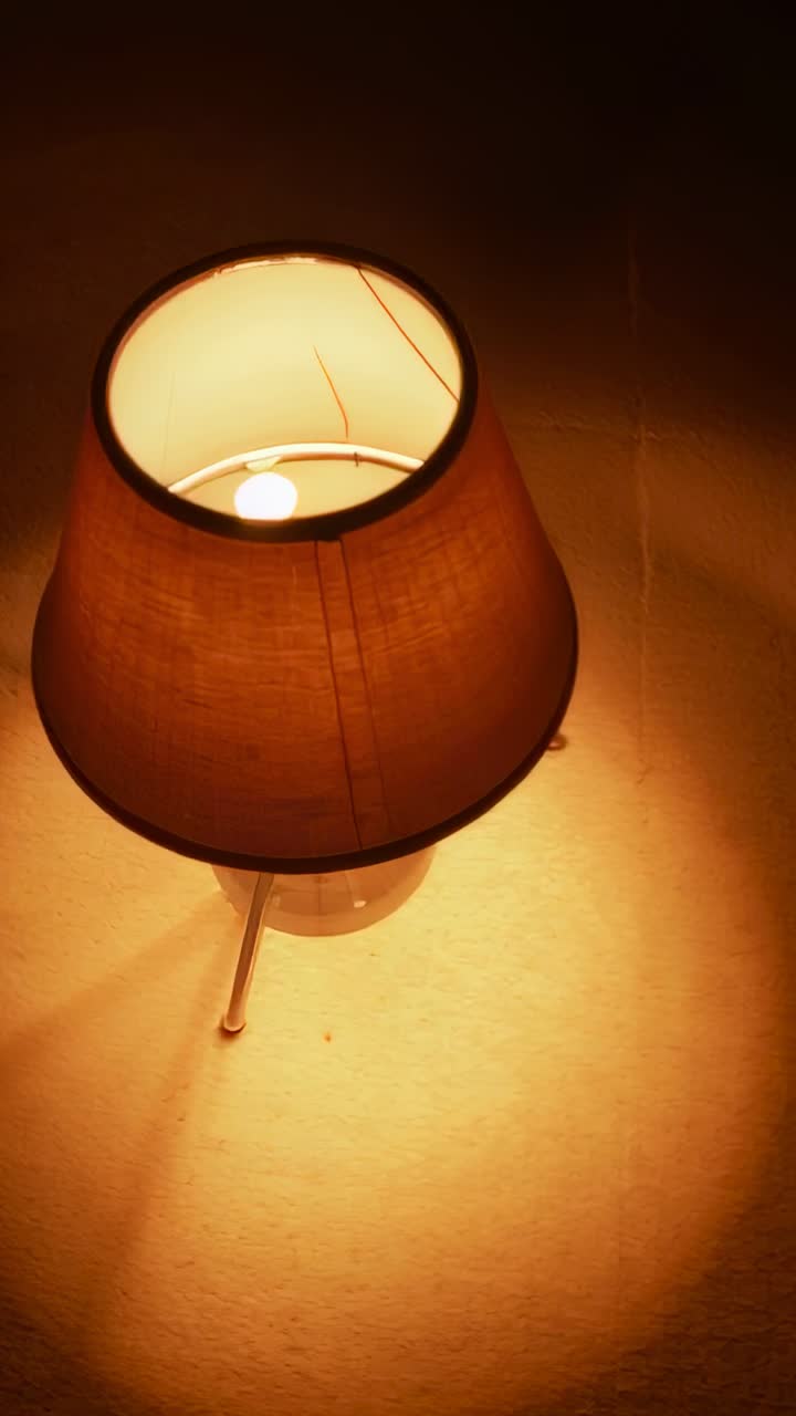 Lampada da tavolo accesa che rivela una pozza di luce calda sul comodino, mostrando lampadina, paralume, base e giuntura della parete