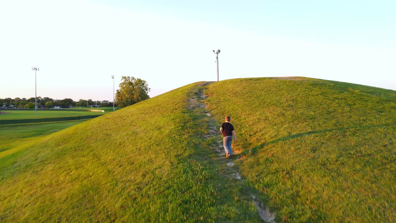 Young boy walking up green hill on sunny evening, aerial follow view
