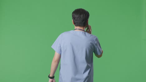 back view of asian male doctor with stethoscope talking on smartphone while walking on green screen background in the hospital