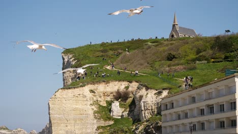 Slow-Motion-of-flying-seagulls-in-front-of-sea-cliffs-and-church-of-Etretat-during-sunny-day---people-walking-uphill-the-mountain-in-background---wide-shot