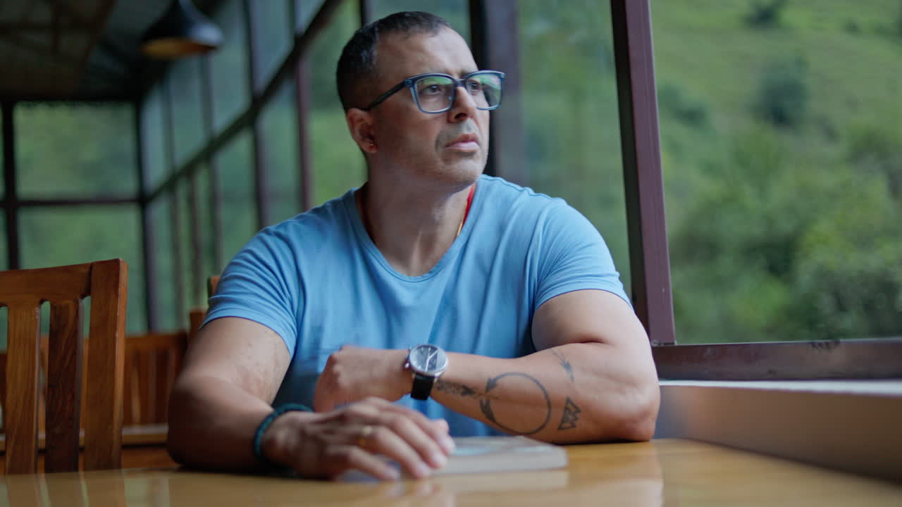 Indian man sitting at cozy corner table with book, gazing thoughtfully out of café window in peaceful countryside setting