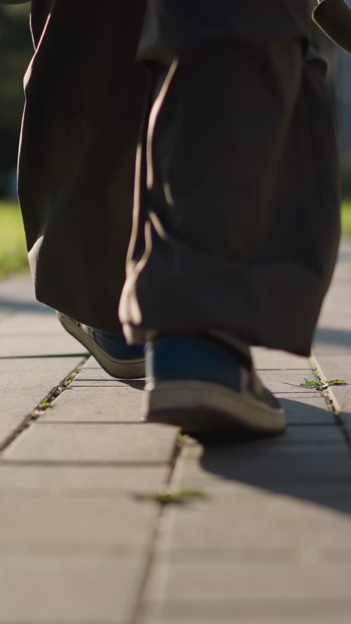 mitad inferior de la persona caminando por un camino pavimentado con pantalones de color oscuro y zapato azul en un pie, ángulo de cámara bajo centrándose en el suelo texturizado, destacando el movimiento casual