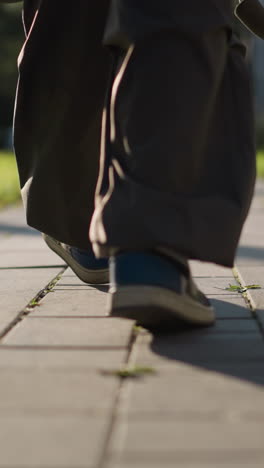 lower half of person walking down paved path wearing dark-colored pants and blue shoe on one foot, low camera angle focusing on textured ground, highlighting casual movement