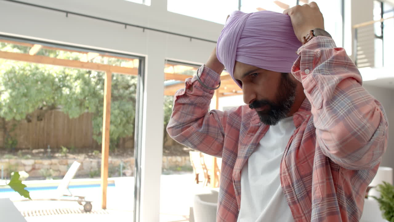 Adjusting turban, man standing indoors in casual attire near window