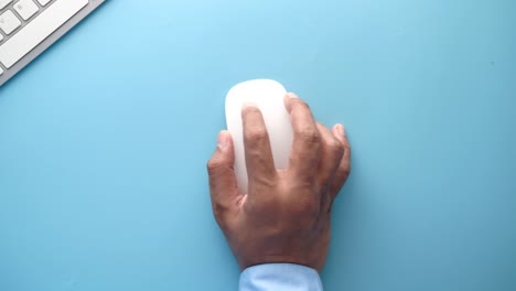 hand using a white computer mouse on a blue desk with a keyboard.