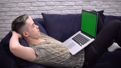 happy young man relaxing on sofa, looking laptop green screen mockup, cozy home